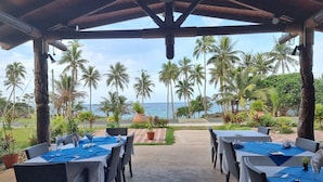 A covered outdoor dining area with tables set for two, overlooking a beach with palm trees and the ocean. - Oasis de Kiamu (Lifou)