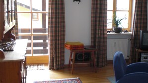 A room with a wooden floor, a window with plaid curtains, a radiator, a table with a lamp, and a chair. - Vacation apartment 1, 68 sqm for 1-2 persons (Berchtesgaden)