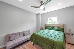 Une chambre avec un lit vert, un canapé gris, un ventilateur de plafond et une fenêtre.