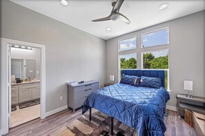 Une chambre à coucher comprenant un lit, une table de chevet, une commode, un ventilateur de plafond et de grandes fenêtres.
