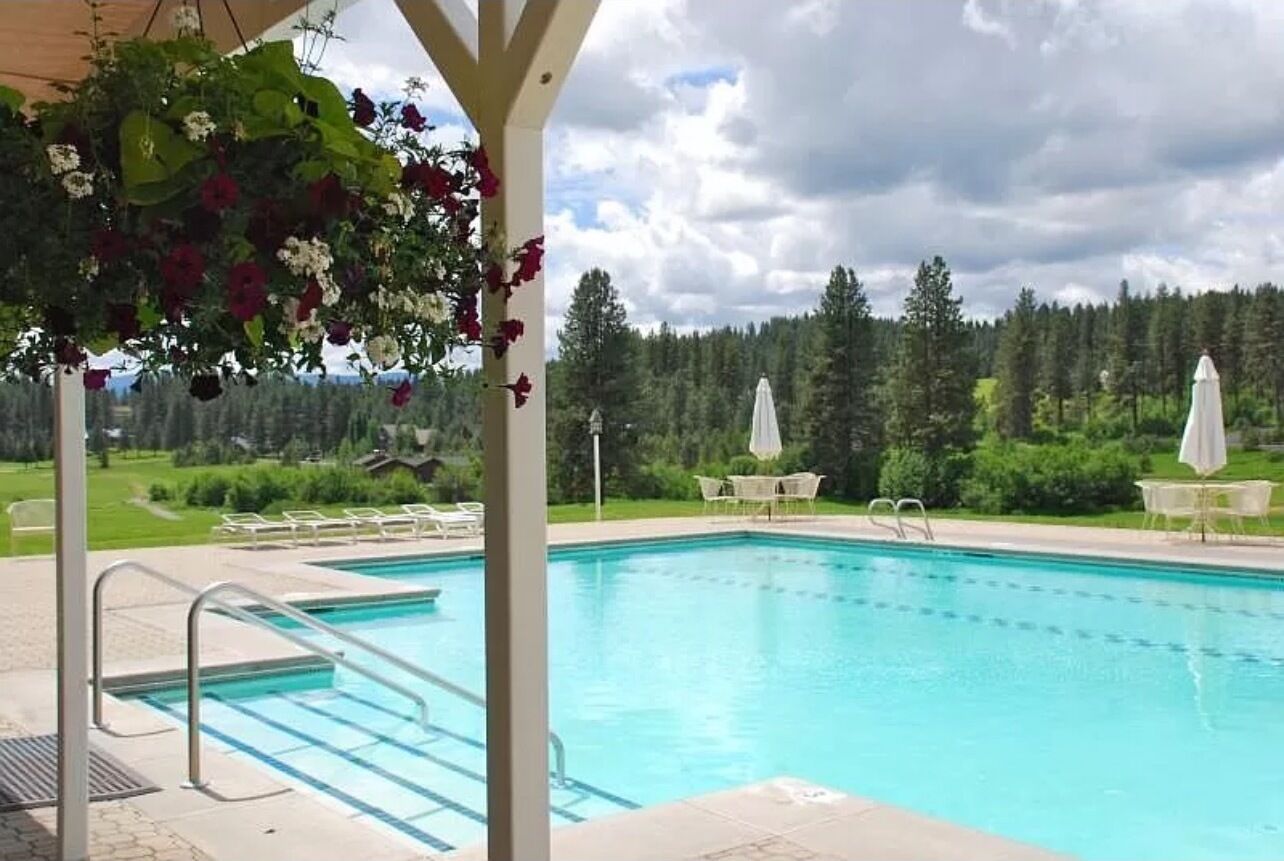 Piscina com vista para uma área arborizada, área de estar ao ar livre e pérgola com flores pendentes.