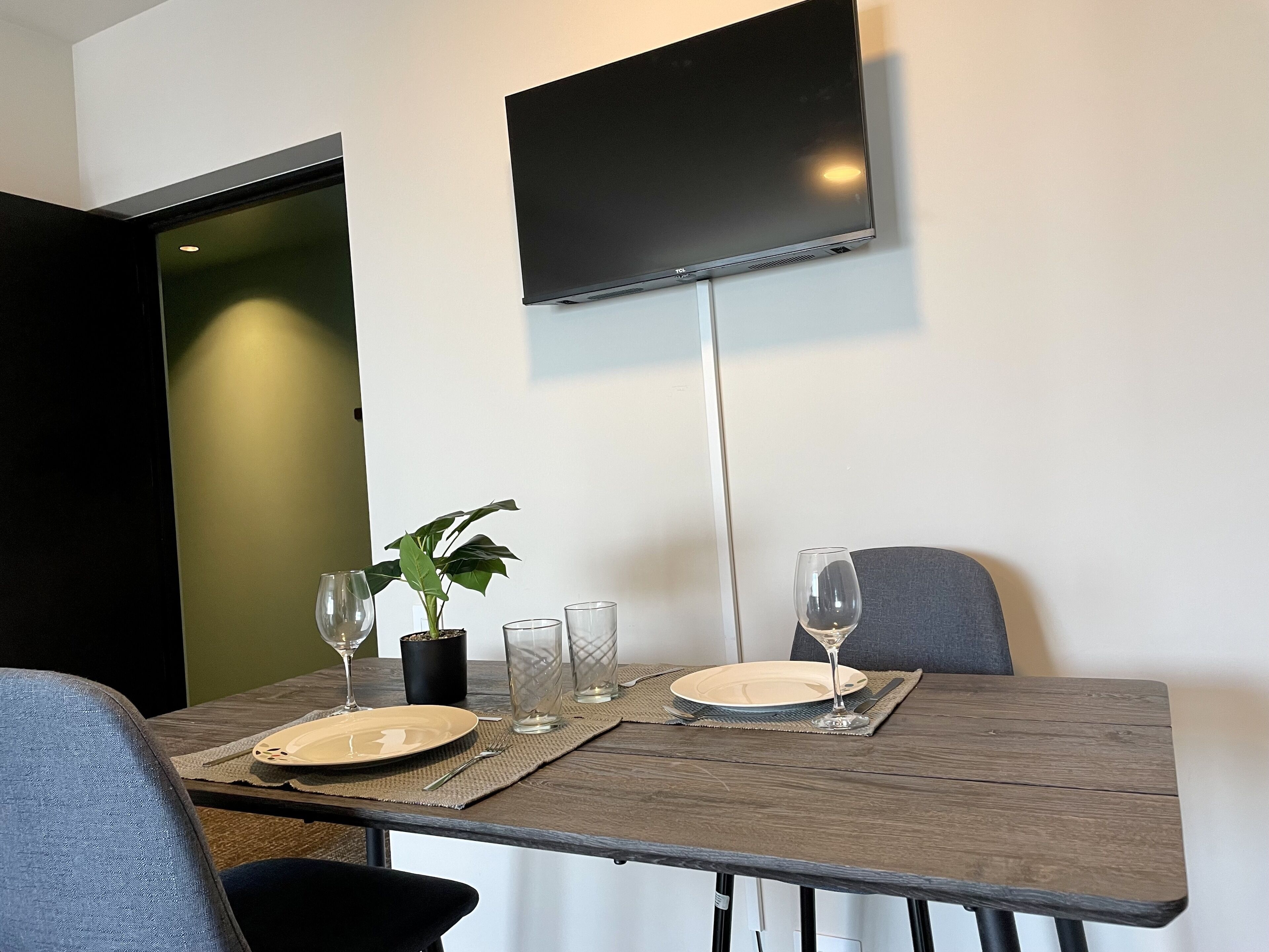 A dining area with a table set for two, a potted plant, and a flat-screen TV mounted on the wall.