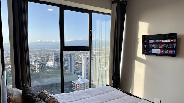 Een slaapkamer met een groot bed, uitzicht op een stadsgezicht door middel van kamerhoge ramen en een tv aan de muur waarop diverse streamingdiensten worden getoond.