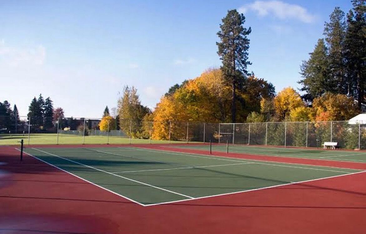 An outdoor tennis court with a red surface, surrounded by trees with autumn foliage.