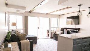 Ein modernes Wohnzimmer mit einer Couch, einem Hocker und einem Couchtisch mit Büchern und einer Vase. Es gibt eine Küchenzeile mit einem Fernseher und Schränken.