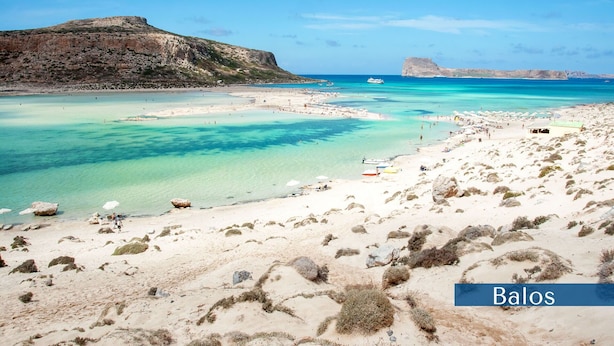Ein Strand mit klarem türkisblauem Wasser, Felsformationen und einem entfernten Gebirge.