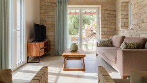 A living room with a stone wall, a wooden TV stand, a beige sofa, and a wooden coffee table. - Villa Jacaranda - Vacation villa with private and heated pool by Agni Bay, Corfu (Agní)
