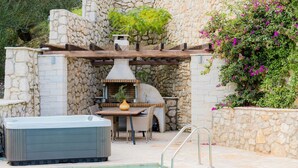 A stone-walled outdoor area with a hot tub, a table with chairs, and a pool. - Villa Jacaranda - Vacation villa with private and heated pool by Agni Bay, Corfu (Agní)