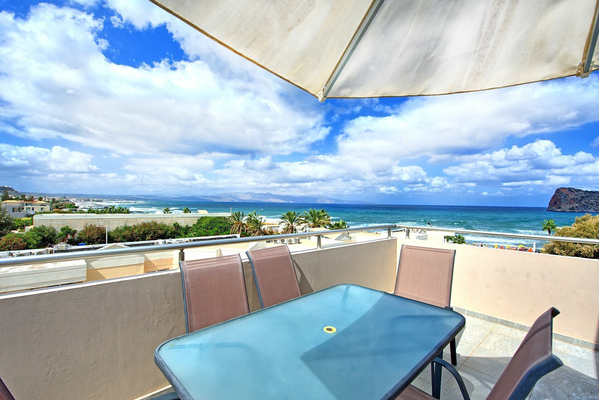 A balcony with a table and chairs, overlooking a beach and ocean with a clear sky.