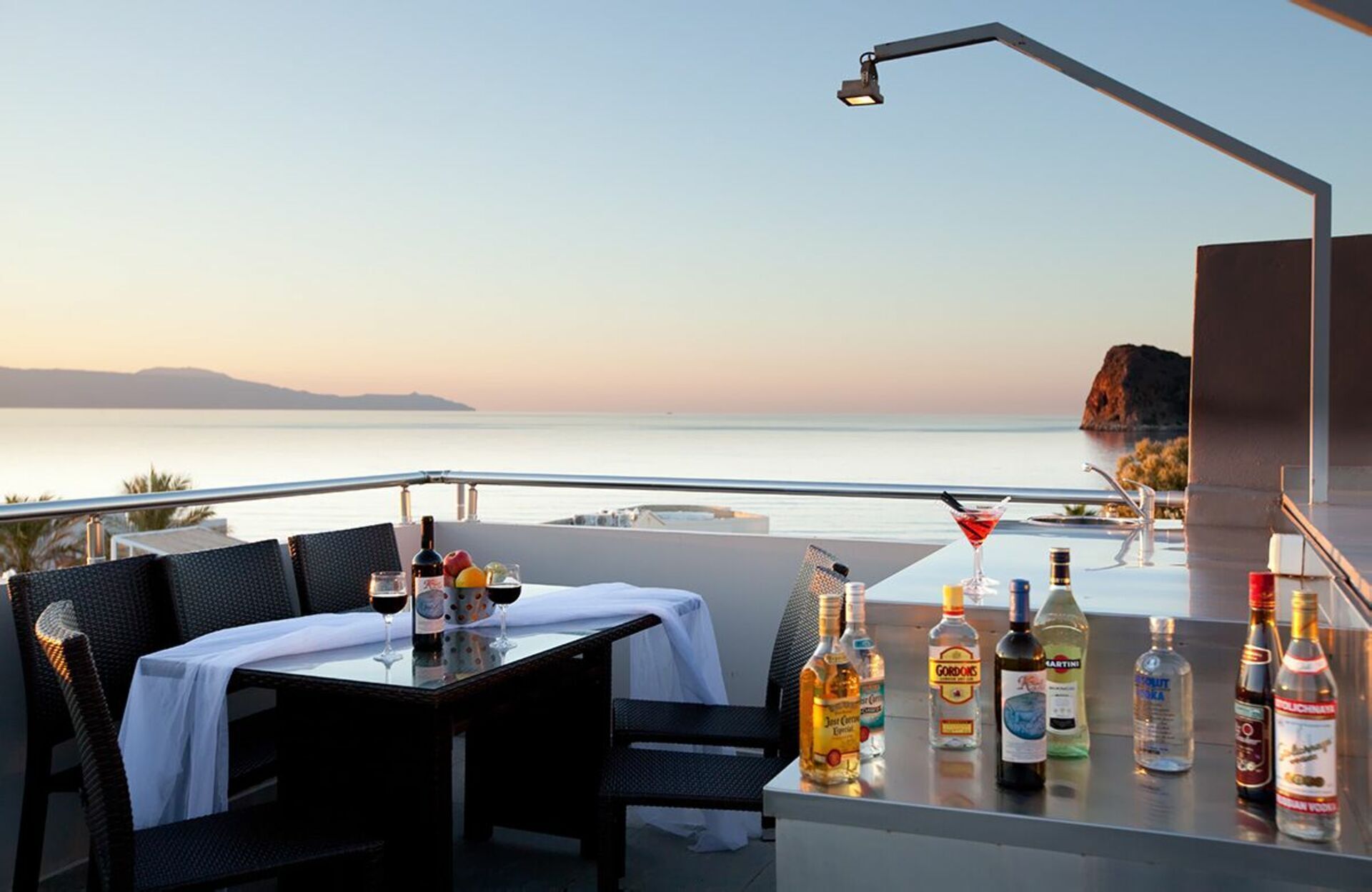 Eine Terrasse mit Tisch für zwei Personen, mit Blick auf ein Gewässer. Auf dem Tisch stehen verschiedene alkoholische Getränke.