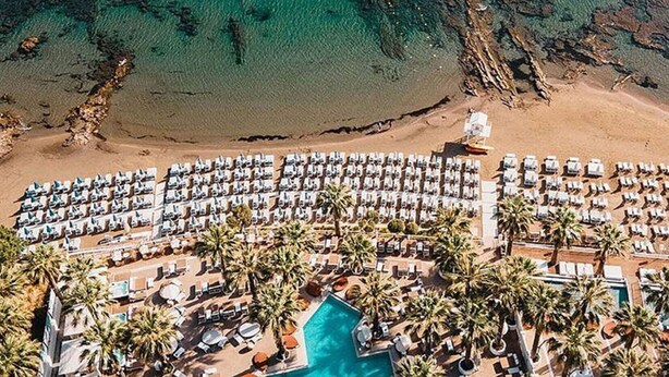 Vista aerea di un resort sulla spiaggia con file di lettini e palme, una piscina e un litorale roccioso.