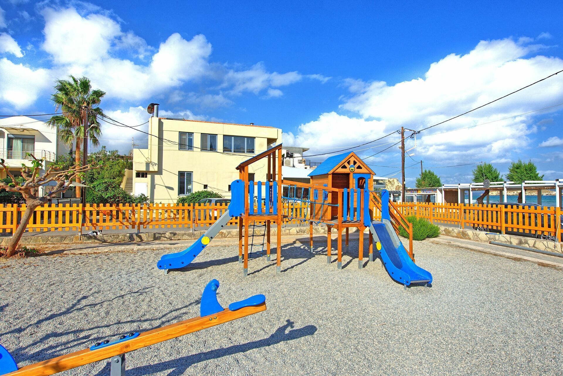 Uma área de recreação infantil com escorrega e estruturas de escalada, cercada por uma grade e edifícios ao fundo.