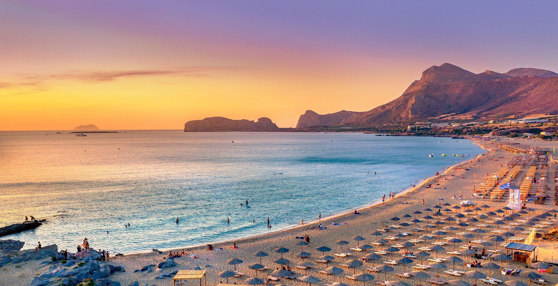 Una playa con sombrillas y tumbonas, cielo despejado y montañas al fondo.