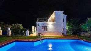 Een modern huis met een zwembad, een balkon en een goed verlichte gevel 's nachts.