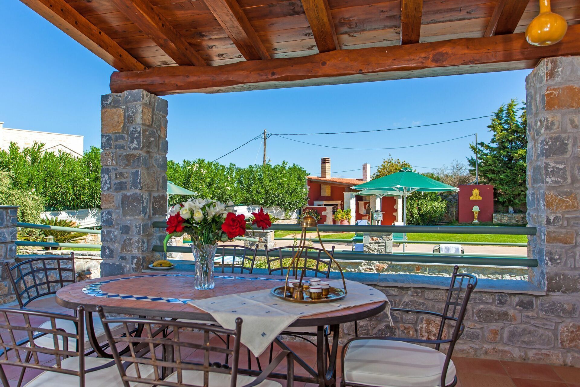 A covered outdoor dining area with stone pillars, a table with chairs, and a view of a building with a green roof.