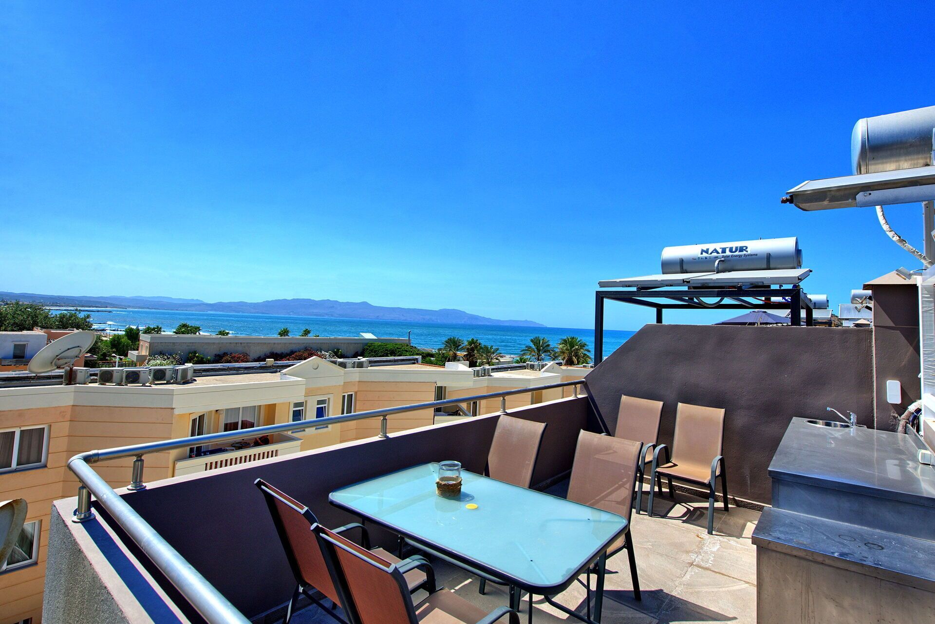 Eine Dachterrasse mit Tisch und Stühlen, Solaranlagen und Blick auf Meer und Berge.