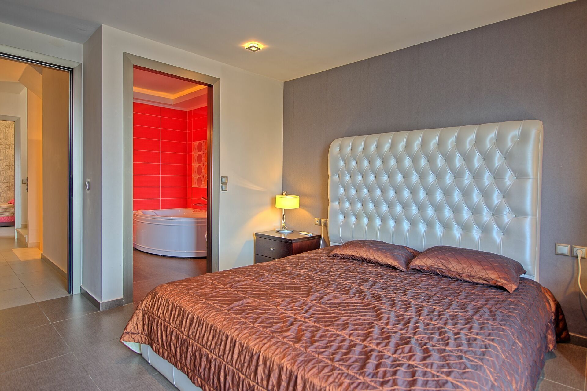 A hotel room with a large bed, a nightstand, a lamp, and a red-tiled bathroom visible through an open door.