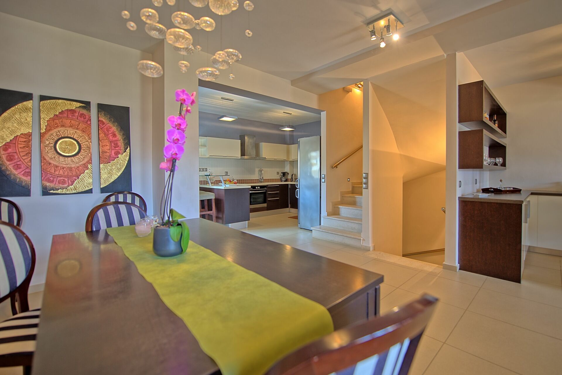 A modern kitchen with a central island, a dining area with a yellow table runner, and a staircase leading to an upper level.
