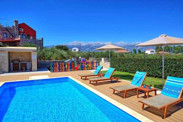 A pool area with lounge chairs and umbrellas, a barbecue setup, and a view of mountains. - Villa Despanto - Vacation villa with pool in Litsarda, Apokoronas, Crete (Litsárdha)
