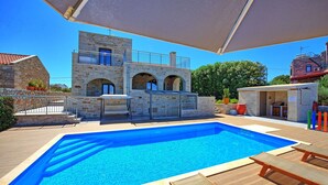 Una piscina con un edificio de piedra al fondo, una zona de estar cubierta y una zona de barbacoa.