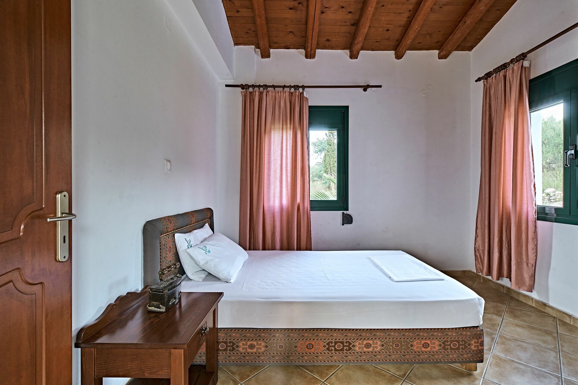 Un dormitorio con techo de madera, una cama con colcha estampada, una mesita de noche y una ventana con cortinas.