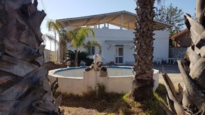 A pool area with a white building in the background, surrounded by palm trees and a stone wall. - Air-conditioned villa - Sea m. 300 (beach) between NOTO-AVOLA (Sr)  (Avola)