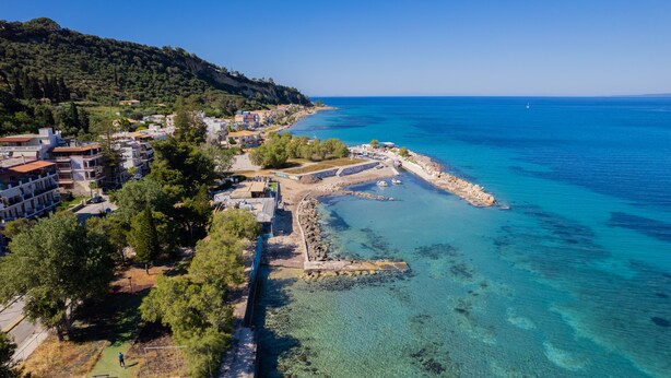 An aerial view of a coastal town with clear blue waters, a rocky breakwater, and buildings along the shore.