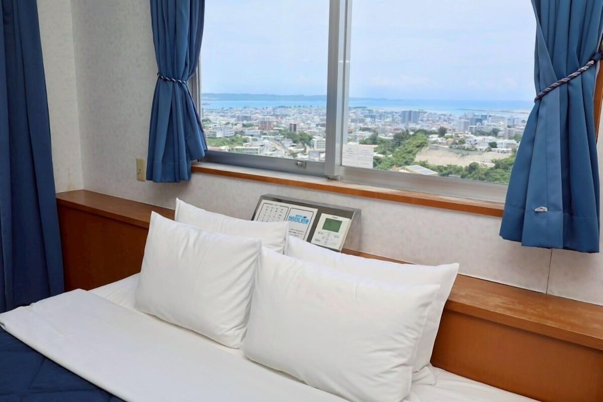 A hotel room with a bed, a window with curtains, and a view of a cityscape.