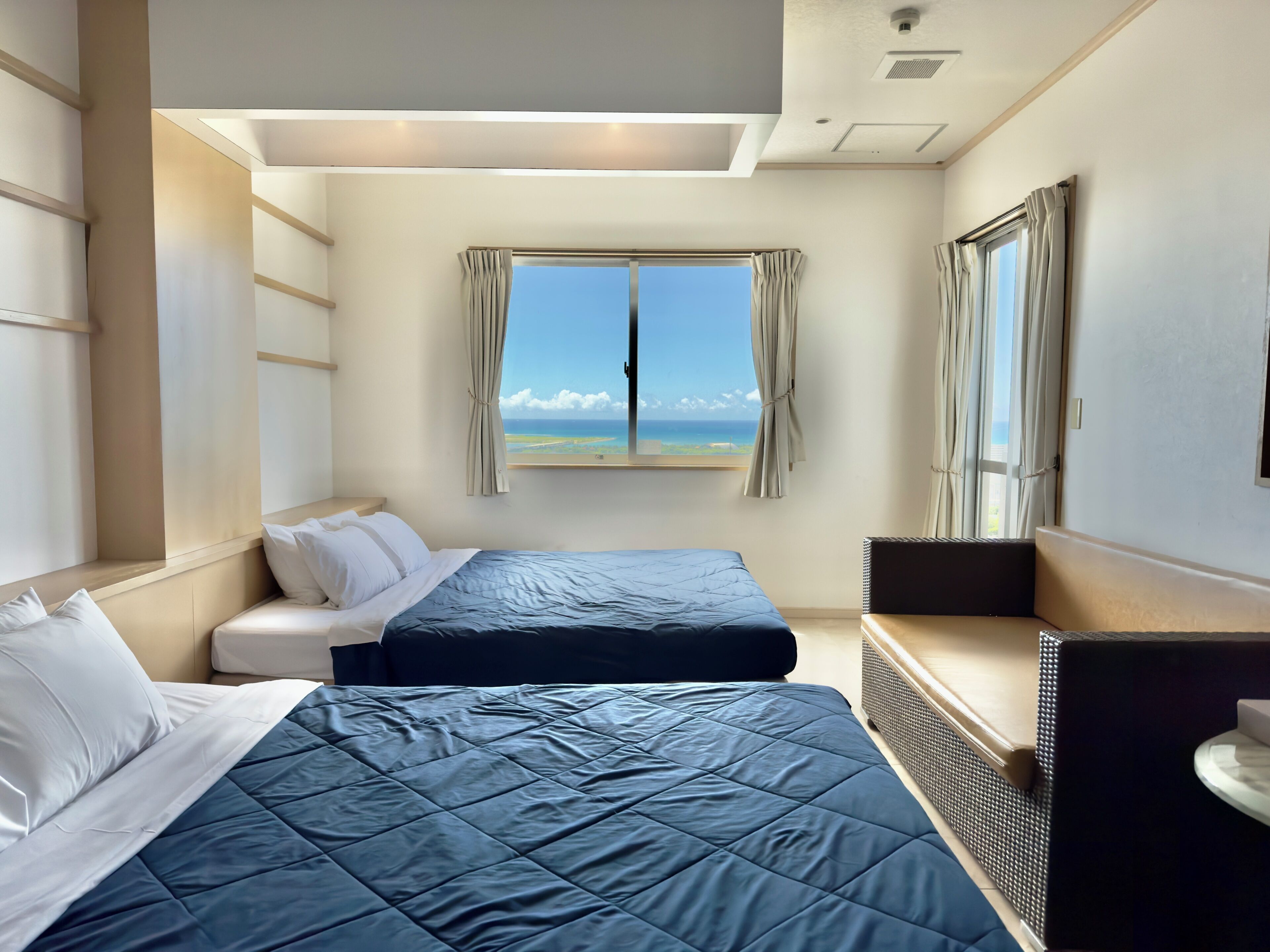 A hotel room with two beds, a large window with curtains, a view of the ocean, and a ceiling with recessed lighting.