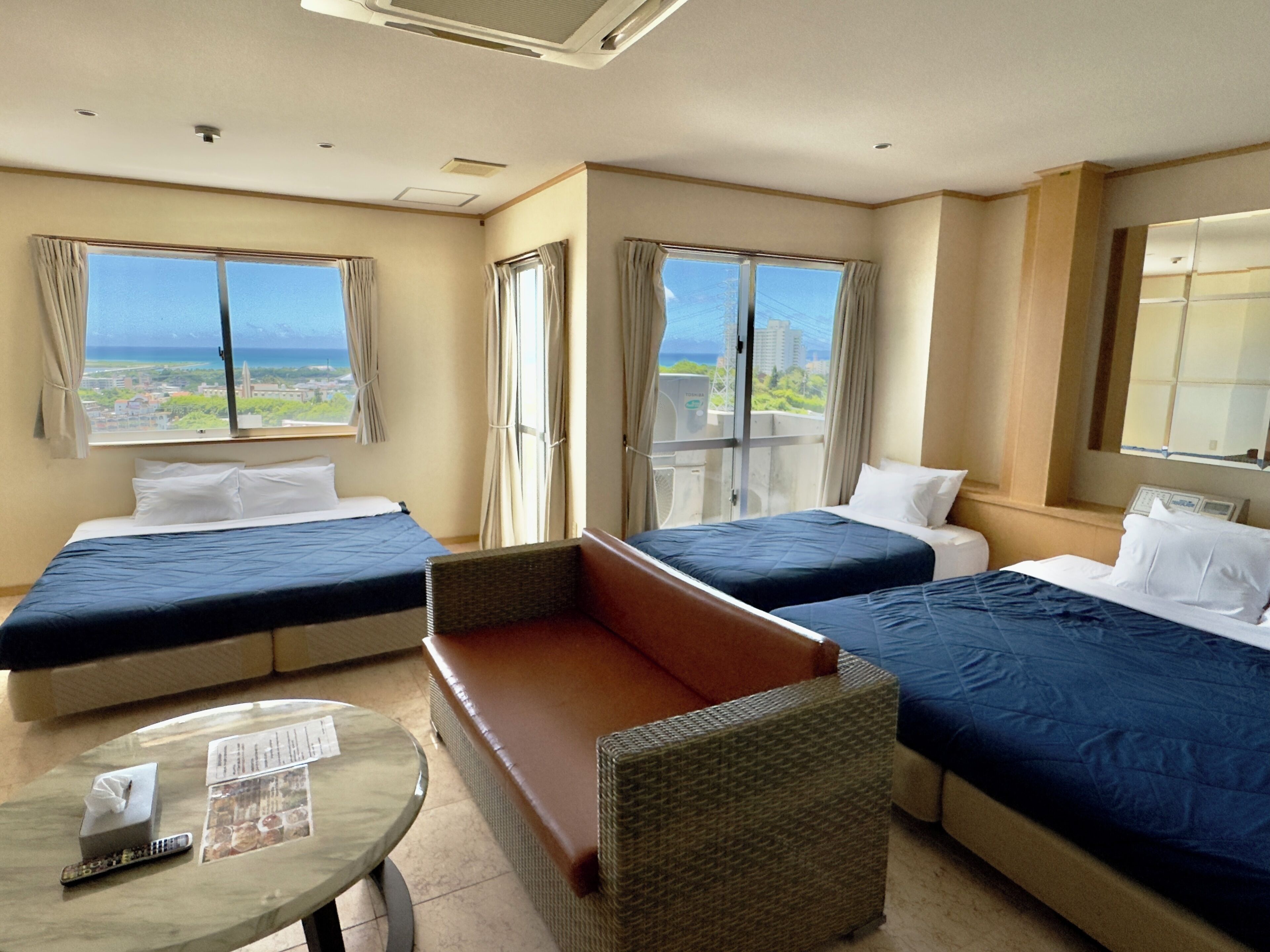A hotel room with two beds, a sofa, a small table with a remote control, and a window with a view of the city.