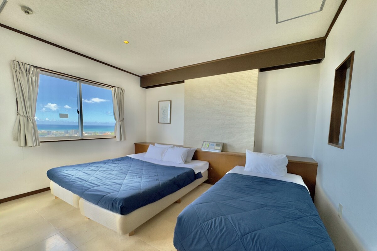 A hotel room with two beds, a window with curtains, a view of the ocean, and a ceiling light.