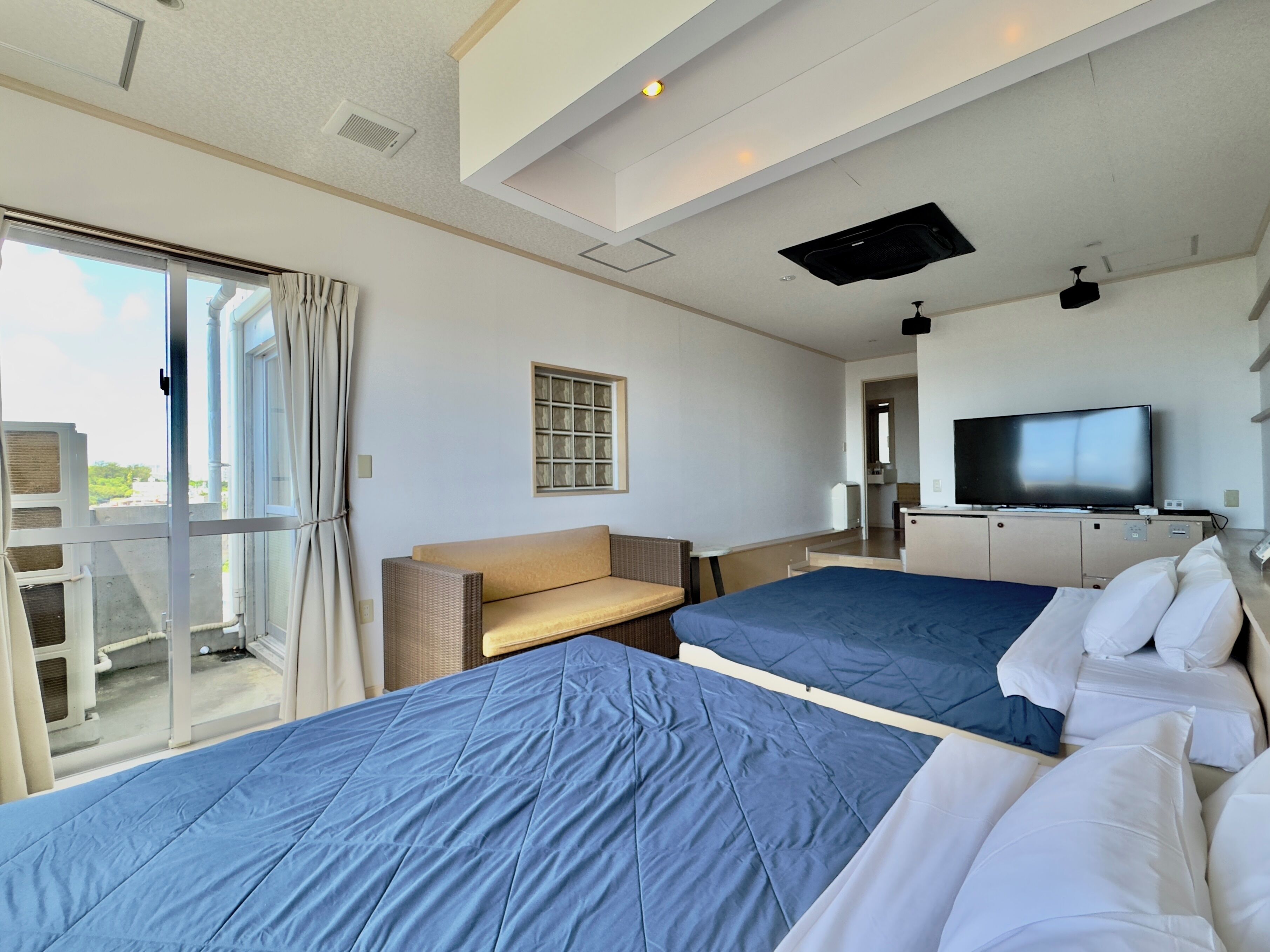 A hotel room with two beds, a television, a window with curtains, and a sliding door to the balcony.