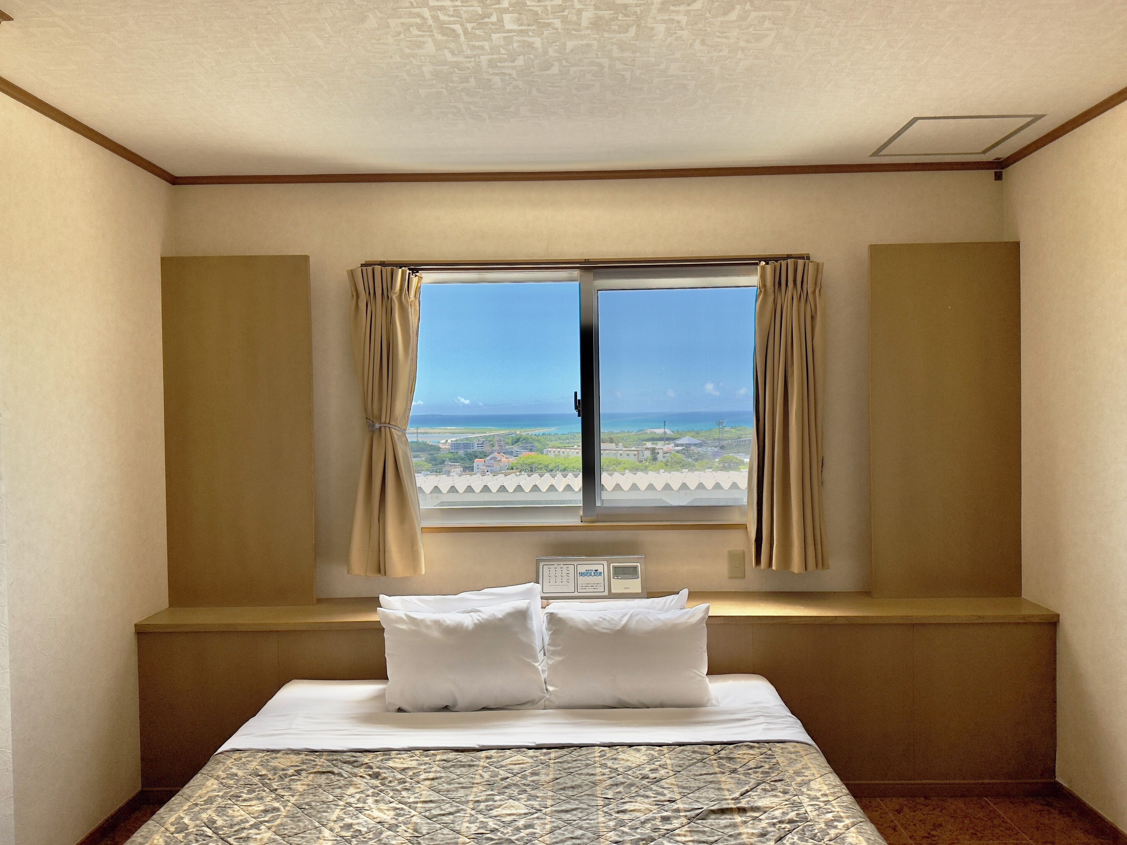 A hotel room with a bed, a window with curtains, a view of the ocean, and a thermostat on the wall.