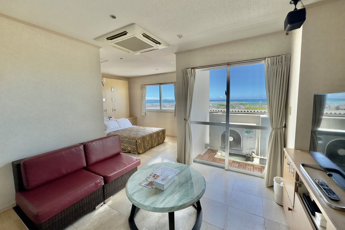 A hotel room with a balcony, a bed, a red sofa, a glass table, and a TV.