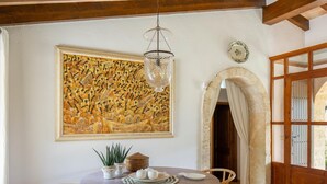 A dining area with a round wooden table, wicker chairs, a hanging light fixture, and a large abstract painting on the wall. - Villa Can Brullet by SunVillas Mallorca (Pollença)