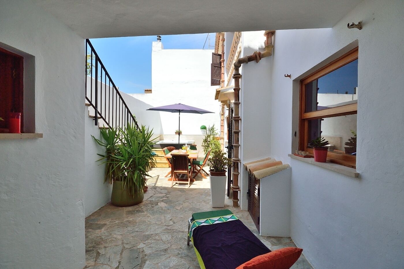 A narrow courtyard with a dining table and chairs, a large potted plant, and a window with a view of the sky.
