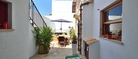A narrow courtyard with a dining table and chairs, a large potted plant, and a window with a view of the sky.