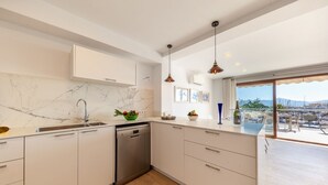 A modern kitchen with marble backsplash, stainless steel appliances, and a view of the marina through large windows. - Apartment Faroles 1 By SunVillas Mallorca (Puerto Pollença)