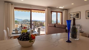 A hotel room with a dining area, a sofa, a large window with a view of the marina, and a table with a bowl of fruit. - Apartment Faroles 1 By SunVillas Mallorca (Puerto Pollença)