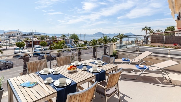 An outdoor dining area with tables set for two, overlooking a marina with boats and a clear sky. - Apartment Faroles 1 By SunVillas Mallorca (Puerto Pollença)