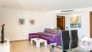 A modern living room with a purple sofa, a glass coffee table, and a painting of a sailboat on the wall. - Apartment Cormes By SunVillas Mallorca (Puerto Pollença)