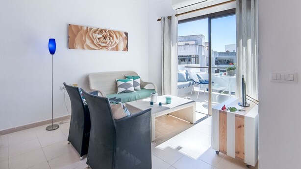 A modern living room with a sofa, a coffee table, and a floor lamp. There is a large window with a view of the city. - Apartment Voltor 2 E By SunVillas Mallorca (Puerto Pollença)