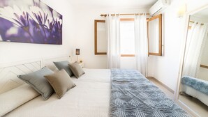 A bedroom with a bed, pillows, a bedside table, a lamp, a window with curtains, and a painting on the wall. - Apartment Voltor 2 E By SunVillas Mallorca (Puerto Pollença)