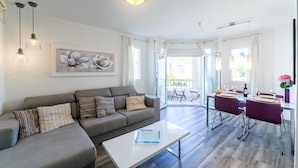 Een moderne woonkamer met een grijze bank, een witte salontafel en een eethoek met paarse stoelen en een glazen tafel.