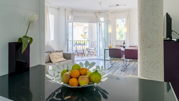 Een moderne keuken met een glazen tafel waarop fruit staat, een eethoek met een tafel en stoelen, en uitzicht op de omgeving door grote ramen.
