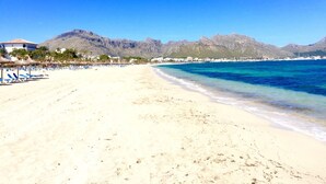 Una playa con aguas azules cristalinas, arena blanca y montañas al fondo.