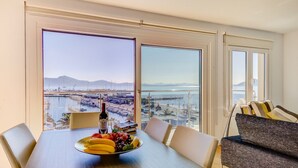 Un comedor con una mesa dispuesta para dos personas, un frutero y una botella de vino. Amplias ventanas ofrecen vistas a un puerto deportivo y montañas distantes.