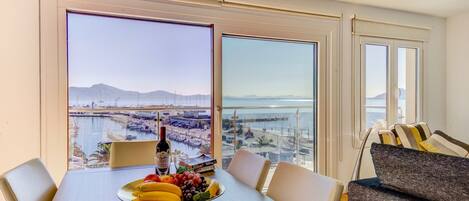 Un comedor con una mesa dispuesta para dos personas, un frutero y una botella de vino. Amplias ventanas ofrecen vistas a un puerto deportivo y montañas distantes.