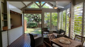 Alpendre coberto com mesa e cadeiras, ventoinha no teto e vista para um jardim através de janelas amplas.