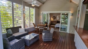 Um terraço espaçoso com móveis de vime, um ventilador de teto e grandes janelas com persianas.